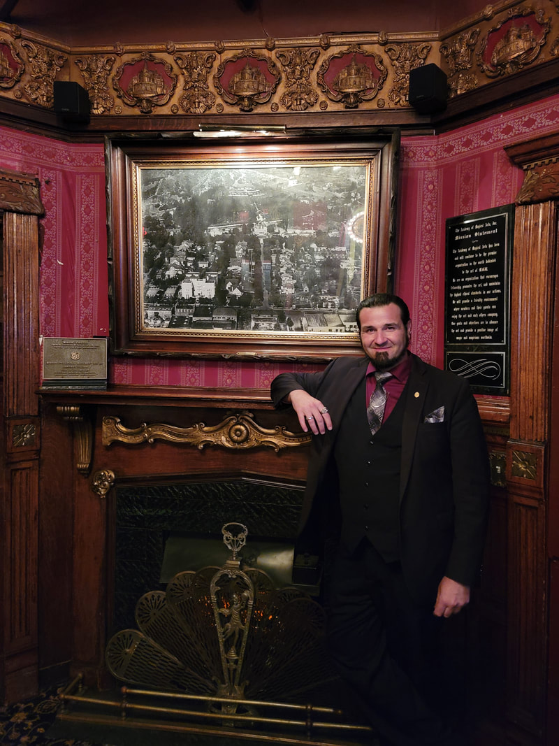 Magician Matthew King at The Magic Castle in Los Angeles standing in front of fireplace in black suit.