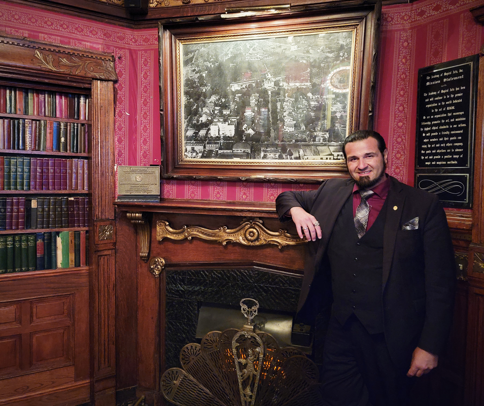 San Diego Magician Matthew King The Magic Castle, wearing a suit in front of fireplace and bookshelf.