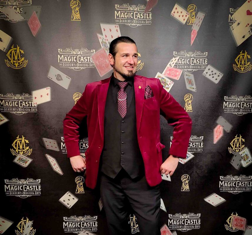 Magician Matthew King in maroon suit at The Magic Castle in Hollywood in front of ornate fireplace and bookcase. 